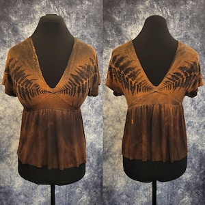 Peut inclure: Un haut à manches courtes avec un décolleté en V profond et une taille empire froncée. Le vêtement présente un effet tie-dye marron et noir avec des motifs de feuilles de fougère sombres partant des épaules vers la poitrine et le dos.