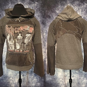 May include: A charcoal gray hooded sweatshirt featuring long sleeves, a front kangaroo pocket, and a distressed graphic print with the text "Under the Demon". The design includes dark fabric patches, decorative metal studs, and visible stitching.