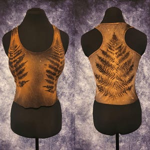 May include: A sleeveless racerback crop top featuring a dark brown and tan bleached effect with a prominent black fern leaf print on the front and back. The garment has a scoop neckline and a fitted silhouette displayed on a mannequin form.