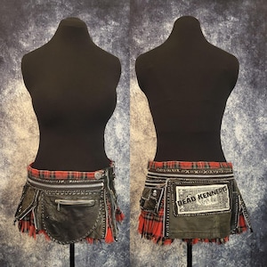 Peut inclure: Mini-jupe en cuir noir et tartan rouge avec plusieurs fermetures éclair et poches. La jupe a un aspect vieilli avec un ourlet effiloché et un patch avec l'inscription "Dead Kennedys Wear". La jupe est présentée sur un mannequin noir.