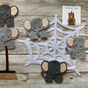 May include: Five grey felt elephant finger puppets with pink ears. The elephants are arranged around a white felt spider web. One elephant is on a brown felt broom stick.
