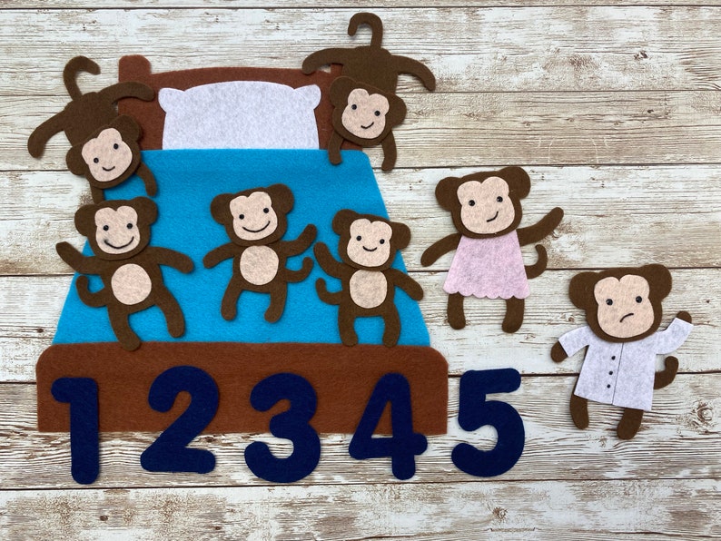 May include: Felt toy set featuring five brown monkeys with different expressions, a blue bed with a white pillow, and the numbers 1 through 5. The monkeys are arranged in a line on the bed, with the last monkey standing off to the side.