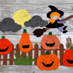 Op de afbeelding: Een set vilten Halloween-decoraties met een heks op een bezemsteel, een gele maan, twee grijze wolken, een bruin hek, groen gras en vijf oranje pompoenen met zwarte gezichten. Een kleine witte kaart toont de tekst Apple Bear Design.