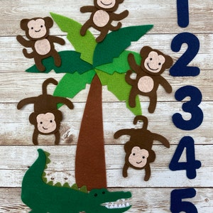 May include: A felt play set with five brown monkeys, a green alligator, and a brown palm tree. The monkeys are climbing the palm tree and the alligator is at the bottom of the tree. There are blue felt numbers 1 through 5 on the right side of the image.