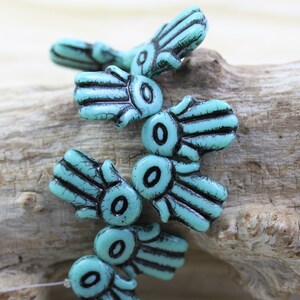 May include: A close-up of turquoise blue hamsa hand beads with black detailing. The beads are arranged in a cluster on a piece of driftwood.