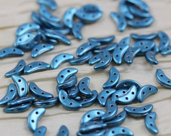 May include: A collection of teal blue crescent-shaped beads with three holes each. The beads are scattered on a white fabric surface.