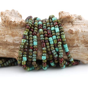 May include: A multi-strand beaded necklace featuring small cylindrical and round beads in shades of turquoise, olive green, and deep maroon. The beads are strung together in several parallel lines, creating a textured appearance with a mix of matte and glossy finishes throughout the strand arrangement.