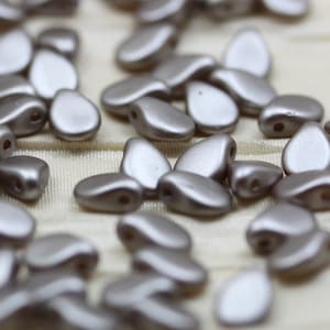 May include: A close-up of a group of silver-colored glass beads. The beads are shaped like teardrops and have a smooth, shiny finish.