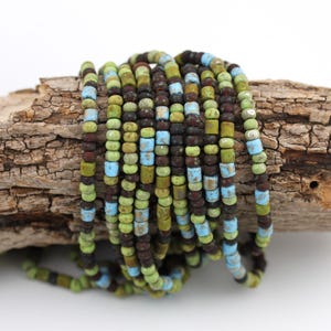 May include: A collection of colourful beads in shades of brown, green, and blue. The cylindrical beads are strung together, creating a textured effect. The beads are resting on a piece of wood.