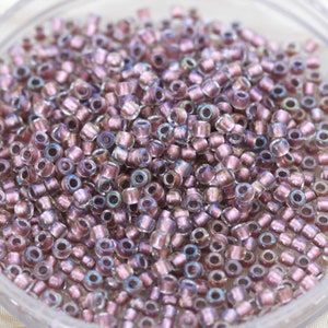 Puede incluir: Primer plano de una gran cantidad de pequeñas cuentas de vidrio redondas e iridiscentes de color morado claro. Las cuentas son translúcidas y tienen un ligero brillo.