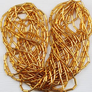 May include: Close-up of a pile of golden, cylindrical beads. The beads are strung together, creating a tangled, textured appearance. The beads are a bright, shiny gold color, reflecting light.