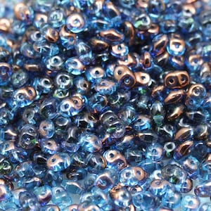 May include: A close-up of a pile of small, iridescent blue and copper glass beads. The beads are shaped like teardrops and have a shiny, reflective surface.