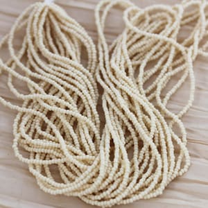 May include: A large quantity of off-white seed beads, arranged in a loose pile. The beads are small and round, and they have a slightly matte finish.