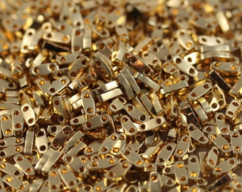 May include: A close-up image of a large quantity of gold-colored metal beads. The beads are small and have a rectangular shape with two holes on each side. The beads are arranged in a random pattern and are piled high in a clear container.