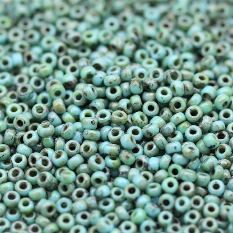 May include: A close-up view of a pile of turquoise blue seed beads. The beads are small and round, with a slightly irregular shape. They have a matte finish and a subtle speckled pattern.