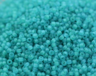 May include: A close-up view of a large quantity of small, round, turquoise-colored glass beads. The beads have a frosted finish and are arranged in a random pattern.