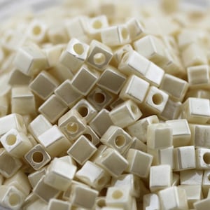 Peut inclure: Un tas de perles carrées ivoire avec une finition nacrée. Chaque perle est percée d'un trou en son centre, parfait pour l'enfilage. Les perles sont éparpillées, créant une surface texturée.