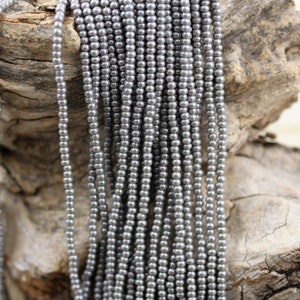 Könnte beinhalten: Eine Schnur silbergrauer Saatperlen mit einem Durchmesser von etwa 2 mm, die vertikal auf einem Holzgrund angeordnet sind.