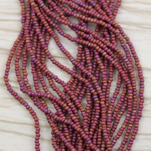 May include: A close-up of a strand of small, round, red and purple seed beads. The beads are arranged in a loose, flowing pattern, creating a textured and colorful effect.
