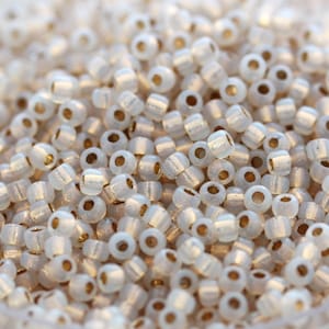 May include: A close-up of a large quantity of small, white, opaque seed beads with a gold-colored hole in the center. The beads have a subtle iridescent sheen.