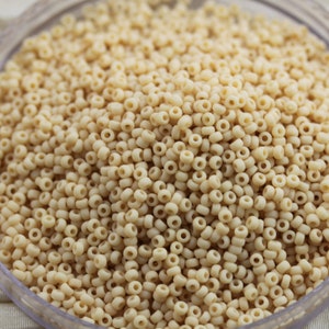以下が含まれることがあります： 小さなオフホワイトのシードビーズの山をクローズアップ。ビーズは均一なサイズと形状で、それぞれの中央に小さな穴があります。それらは密集して詰め込まれ、フレームを埋めています。