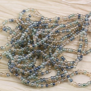 May include: A strand of seed beads in shades of beige, gray, and blue. The beads are small and round, and they are strung together on a clear thread.