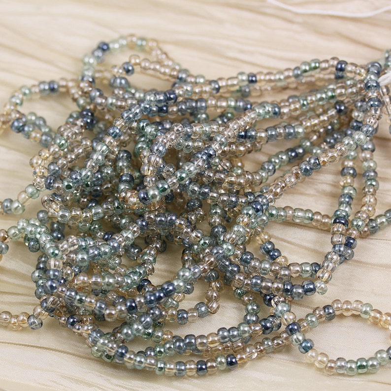 May include: A strand of seed beads in shades of beige, gray, and blue. The beads are small and round, and they are strung together on a clear thread.