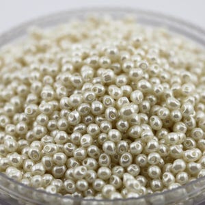 May include: A close-up shot of a container filled with ivory-colored, round beads. The beads have a pearlescent sheen, suggesting they are made of faux pearl material. The beads are densely packed, filling the frame.