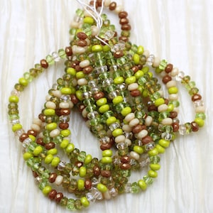 May include: A collection of beaded strands in green, brown, and cream hues. The beads are strung together, forming a textured, layered design. These beads are likely for jewellery making or crafting projects.