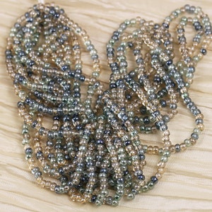 May include: A strand of seed beads in shades of brown, gray, and clear. The beads are small and round, and they are strung together in a long, continuous strand.