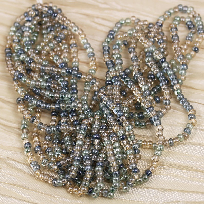 May include: A strand of seed beads in shades of brown, gray, and clear. The beads are small and round, and they are strung together in a long, continuous strand.