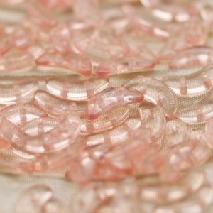 May include: A close-up of a pile of pink, translucent, crescent-shaped beads. The beads are made of glass and have a smooth, shiny finish.