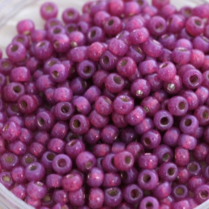 May include: A close-up image of a pile of small, round, purple glass beads with a metallic sheen. The beads have a slightly textured surface.