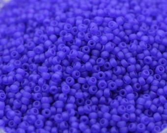 May include: A close-up view of a large quantity of small, round, opaque purple seed beads. The beads are arranged in a random pattern, creating a textured surface.