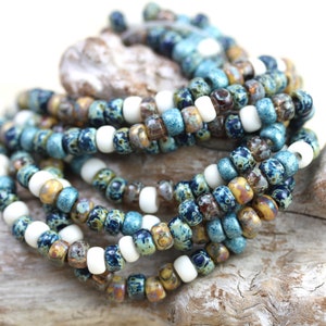 May include: A close-up of a strand of blue, white, and brown seed beads. The beads are arranged in a random pattern and are strung on a clear thread.