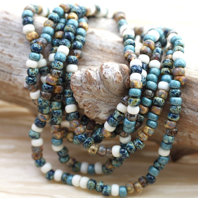 May include: A close-up of a multi-strand necklace made of small, colorful glass beads. The beads are in shades of blue, brown, and white, and they are arranged in a random pattern. The necklace is lying on a piece of driftwood.