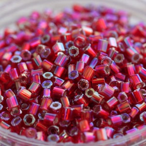 May include: A close-up of a pile of small, red, iridescent glass beads. The beads are cylindrical in shape and have a slightly metallic sheen.