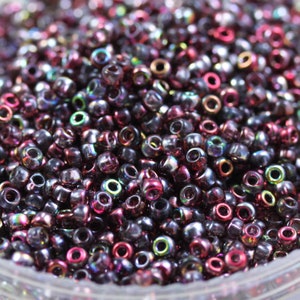 May include: A close-up of a large quantity of small, round, iridescent beads. The beads are a mix of dark purple, silver, and gold colors.