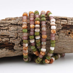 May include: Several beaded bracelets in shades of green, white, brown, and purple. The bracelets are arranged on a piece of textured wood, giving a natural feel. The small, round beads create a colourful and tactile accessory.