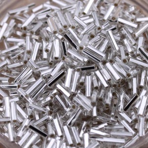 May include: A close-up shot of a pile of silver-colored, cylindrical beads. The beads are small, with a reflective, metallic finish. Each bead has a small, dark band near the center. The beads are scattered, creating a textured surface.