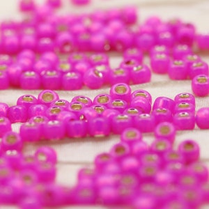 Puede incluir: Primer plano de un montón de pequeñas cuentas de vidrio rosa iridiscente. Las cuentas tienen un acabado metálico plateado.