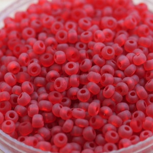 May include: A close-up view of a bowl filled with small, opaque, red glass beads. The beads are round and have a slightly frosted finish.