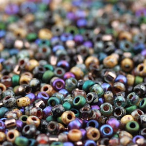 May include: A close-up of a collection of small, multi-coloured seed beads. The beads are a mix of green, purple, gold, and black. Each bead is round with a central hole, ideal for jewellery making and crafts.