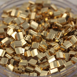 Puede incluir: Primer plano de un recipiente lleno de cuentas de metal de color dorado. Las cuentas son pequeñas y de forma cuadrada con dos agujeros en cada cuenta.
