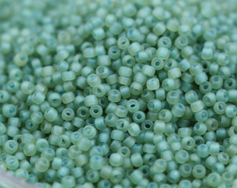 May include: A close-up view of a large quantity of small, round, light green glass beads. The beads have a frosted finish and are arranged in a random pattern.