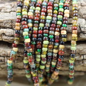 May include: A strand of colourful seed beads in various shades of green, brown, red, and cream. The beads are small and round, and they are arranged in a random pattern.