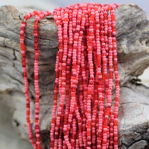 May include: A strand of red and pink seed beads, arranged in a cascading pattern, against a rustic wooden background.