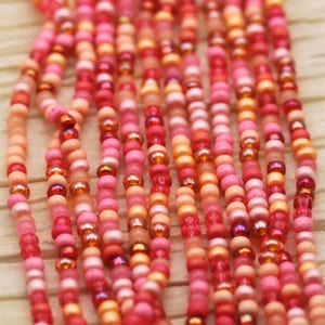 Puede incluir: Primer plano de una hebra de cuentas rosadas, naranjas y rojas. Las cuentas son pequeñas y redondas, y están enhebradas juntas en un patrón aleatorio.