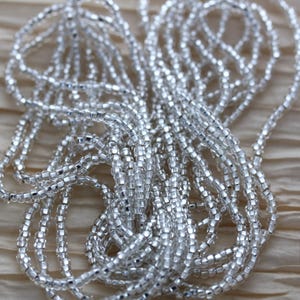 May include: Close-up of multiple strands of silver-colored beads. The beads are small, square-shaped, and strung together, creating a shimmering effect. The background is a light, neutral color, providing contrast to the beads. The beads appear to be a decorative item.