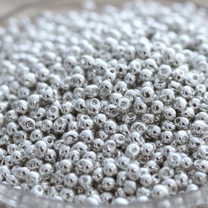 May include: A close-up of a large quantity of silver seed beads. The beads are small and round, with a slightly elongated shape. They are all the same size and have a shiny, metallic finish.